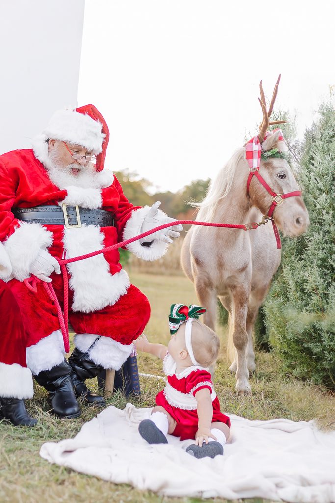 Santa Claus sitting beside Burt the Reindeer and a baby dressed for Christmas on a white blanket during a Birmingham, Alabama Christmas mini session.