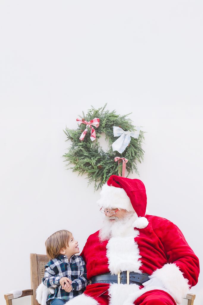A young boy in a blue plaid shirt smiling up at Santa Claus beneath a Christmas wreath during a holiday portrait session with 5U Photography.