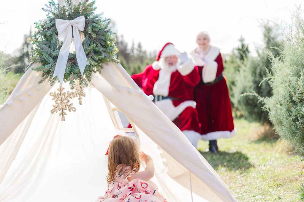 A little girl sitting beside a Christmas teepee as Santa Claus and Mrs. Claus wave to her during a Christmas mini session near Birmingham, Alabama.