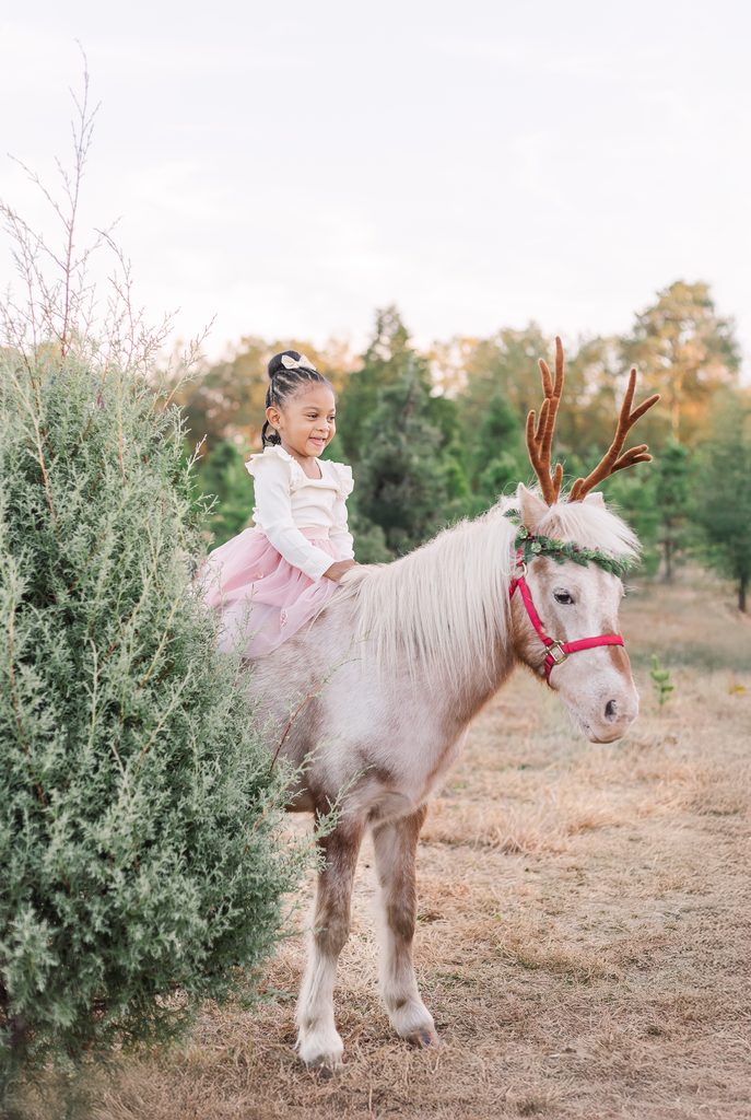 A smiling little girl in a pink tutu riding Burt the Reindeer among Christmas trees during a holiday mini session near Birmingham, Alabama.