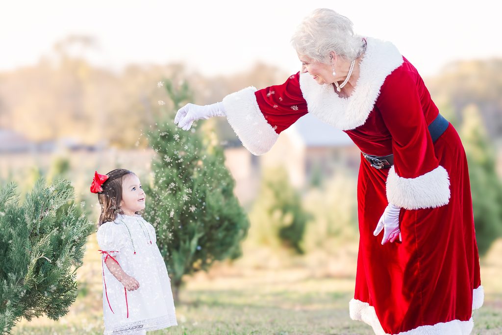 Mrs. Claus reaching toward a smiling little girl in a white dress during an outdoor Christmas mini session among evergreen trees near Birmingham, Alabama.