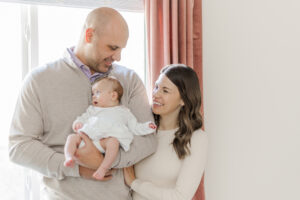 Mom and dad standing in front of window holding newborn baby girl during milestone membership in-home newborn session at their home in Birmingham, AL