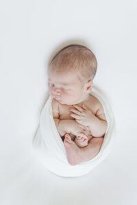 newborn baby boy laying on white blanket swaddled in white swaddle during in-home, lifestyle newborn session as a part of milestone membership