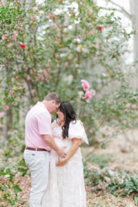 Father and mother lovingly embrace in front of a blooming pink peony bush, surrounded by lush greenery during spring maternity session at the birmingham botanical gardens