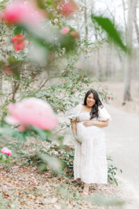 Expecting mother smiling in front of a blooming pink peony bush at Birmingham Botanical Gardens, gently holding her baby bump