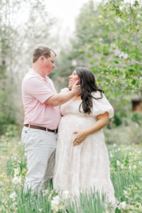 mom and dad standing with dad's hand on moms cheek looking down at her lovingly, with mother’s hands on her baby bump, with tulips surrounding her feet during spring maternity session