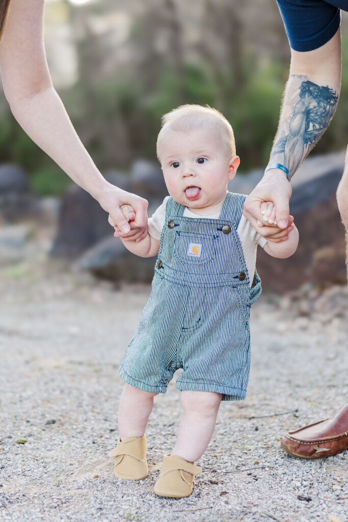 6 month old baby boy standing on gravel path helped by mom and dad holding each hand