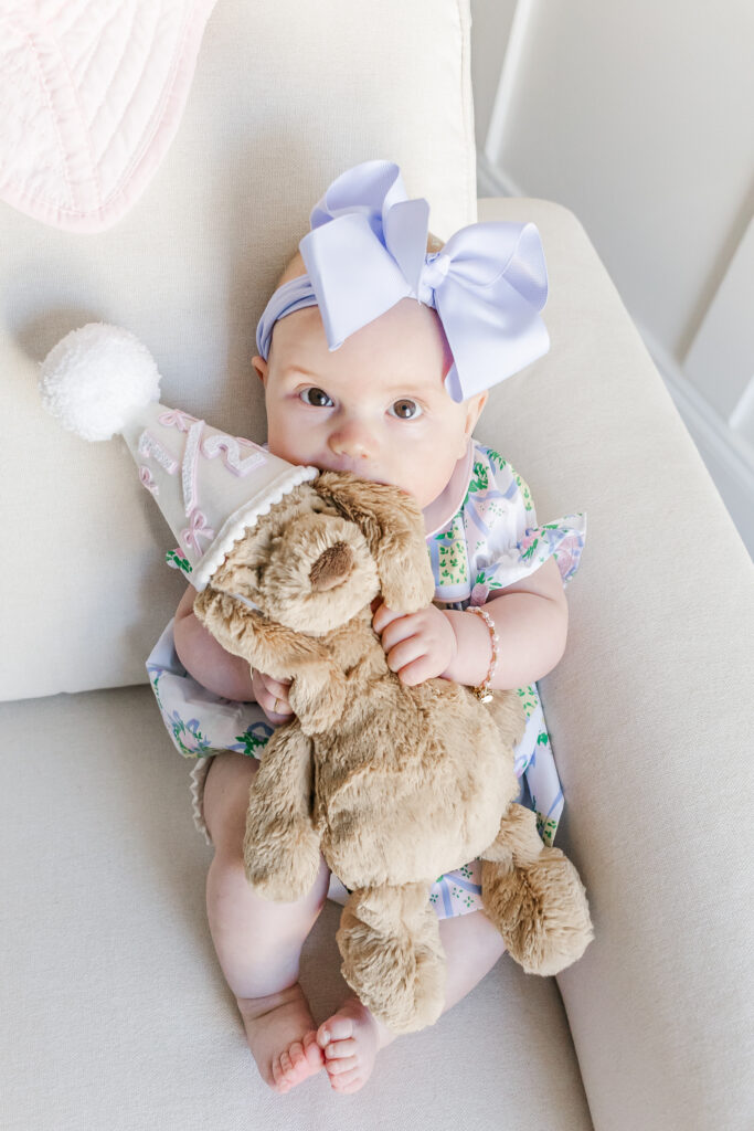 Ella celebrating her 6-month milestone in a soft pink and white nursery filled with love and light holding stuffed puppy wearing party hat with 1/2 on it for her 1/2 birthday
