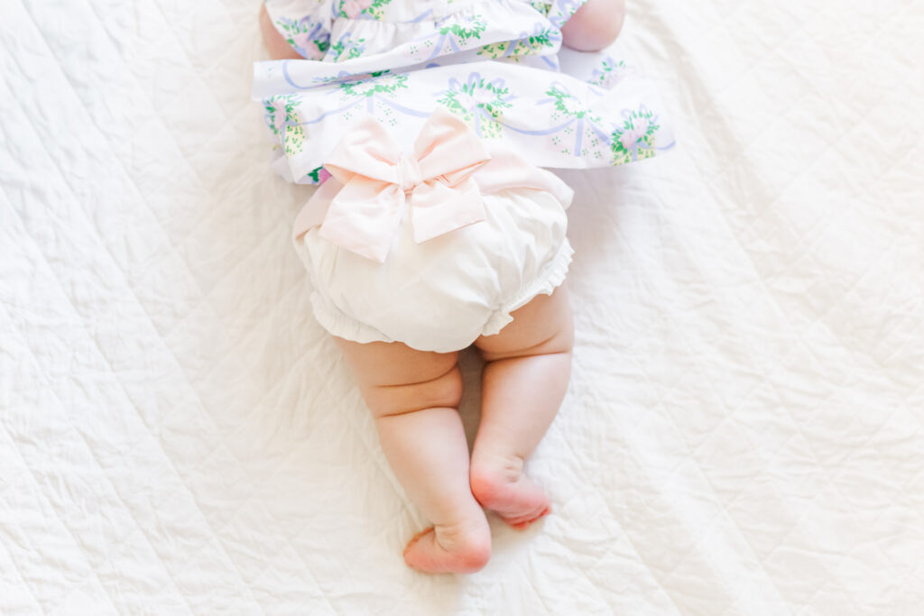 Close up of beaufort bonnet company's white ruffled diaper cover with pink bow while Ella lays on her stomach on white blanket