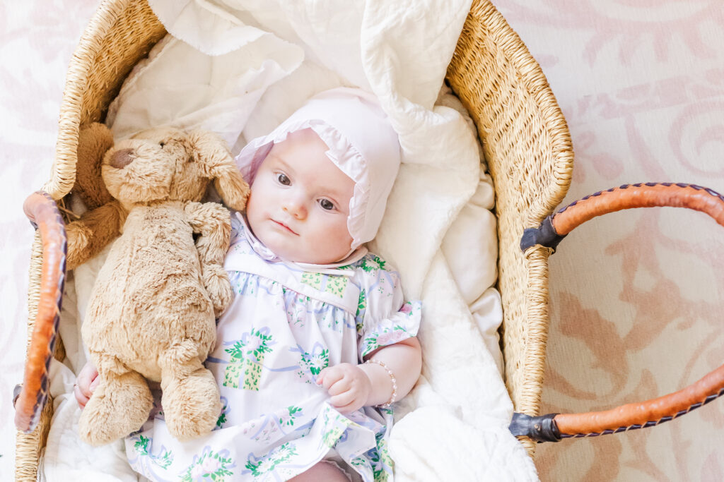 Little girl laying in wicker moses basket wearing a pink ruffled bonnet holding Jelly cat stuffed dog 