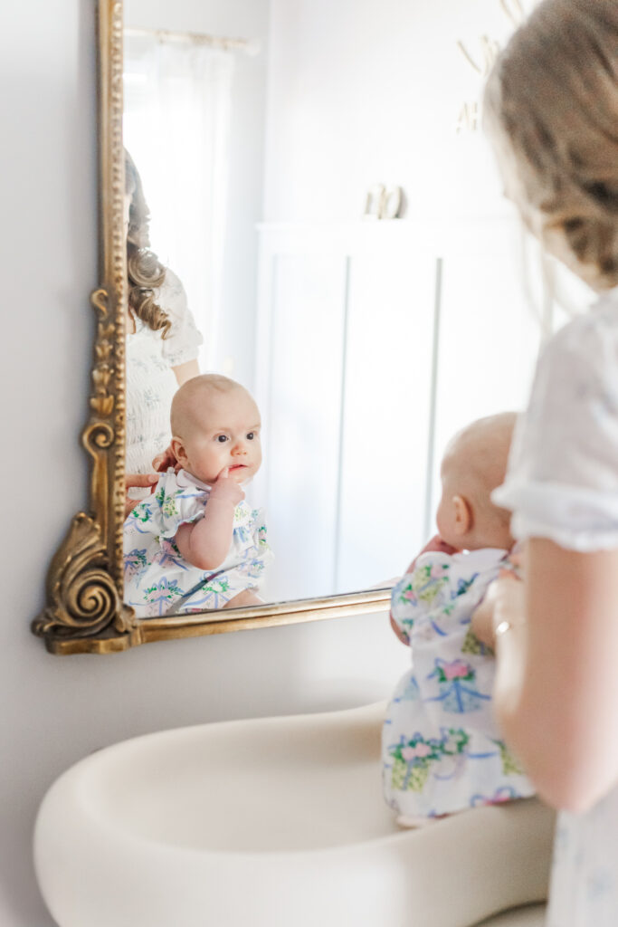 picture of Little girl's reflection as she looks in the mirror while mom buttons her beaufort bonnet company dress getting ready for her milestone membership session celebrating 6 months