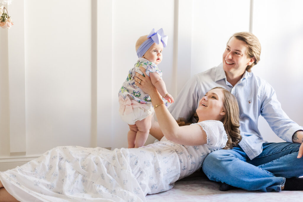 Mom laying on nursery rug with head resting on dad's knee holding 6 month old Ella up while she smilies at the camera during 5U Photography's milestone session