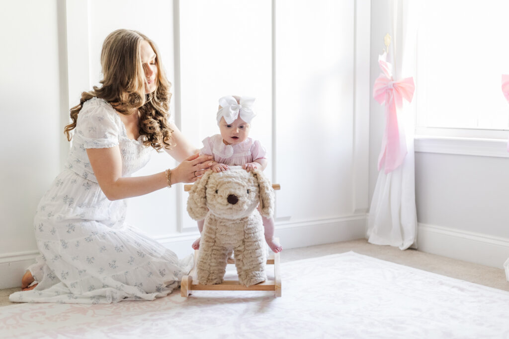 Mom kneeling on pastel pink nursery rug helping 6 month old daughter sit on plush rocking horse in pink dress during 6 month milestone session 