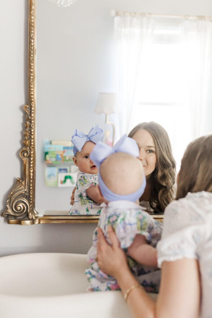 Mom holding 6 month old little girl while she sits on changing table looking in the mirror smiling at her reflection during 6 month session with 5U Photography