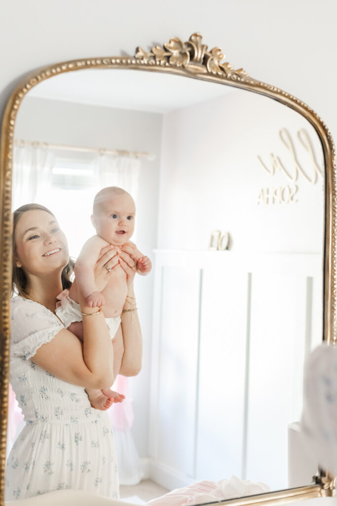 Mom holding up little girl in her diaper looking in mirror smiling joyfully in the nursery at their home in calera alabama
