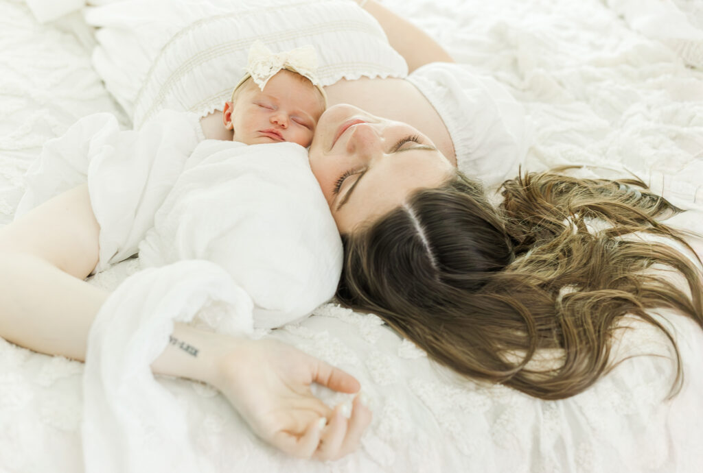 Newborn baby Eleanor swaddled in a soft pink blanket, sleeping peacefully on a white bed in a bright, airy bedroom cradled by mom