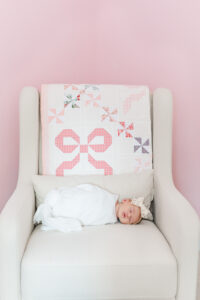 Newborn Eleanor swaddled in white swaddle with matching bow laying in rocker in pink nursery with hand made bow quilt folded over back during in-home newborn session at family's home in highland lakes