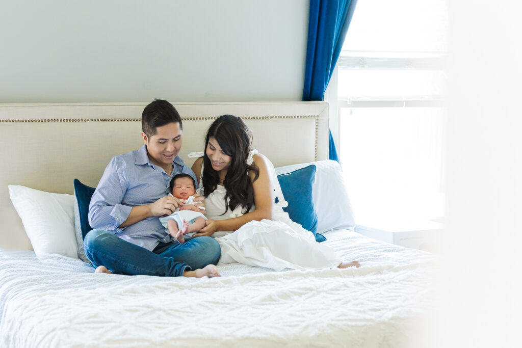 Parents sitting on bed with newborn baby boy in their arms, smiling together during a Trussville, Alabama lifestyle newborn session.