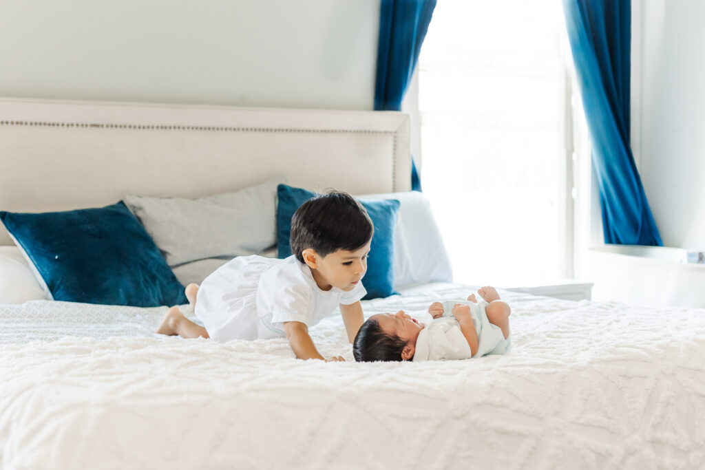 Toddler big brother leaning close to newborn baby brother on a white bed during an in-home newborn session in Trussville, Alabama.