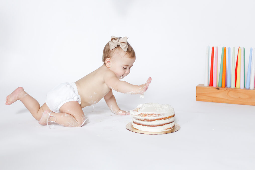 Greystone In-home one year milestone cake smash session in Birmingham, Alabama, featuring baby girl reaching for her birthday cake on a white backdrop.