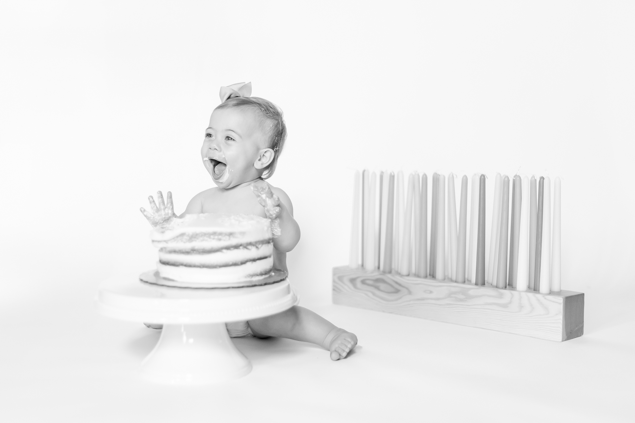 Black and white one year milestone cake smash portrait of baby girl laughing with joyful expression beside a simple cake on a white pedestal.