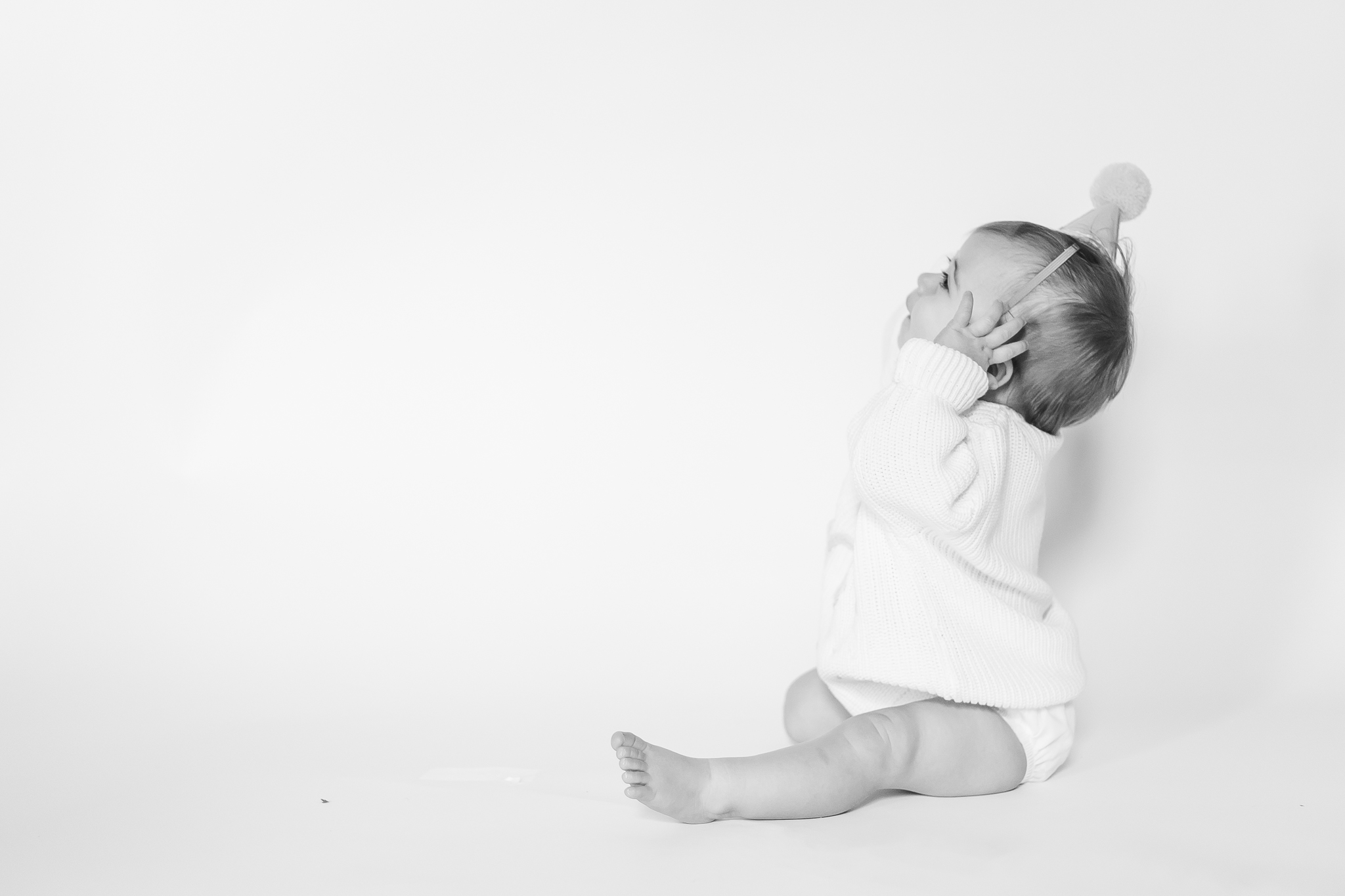 Black and white one year milestone portrait of baby girl wearing a simple first birthday hat, captured on a minimalist white backdrop.