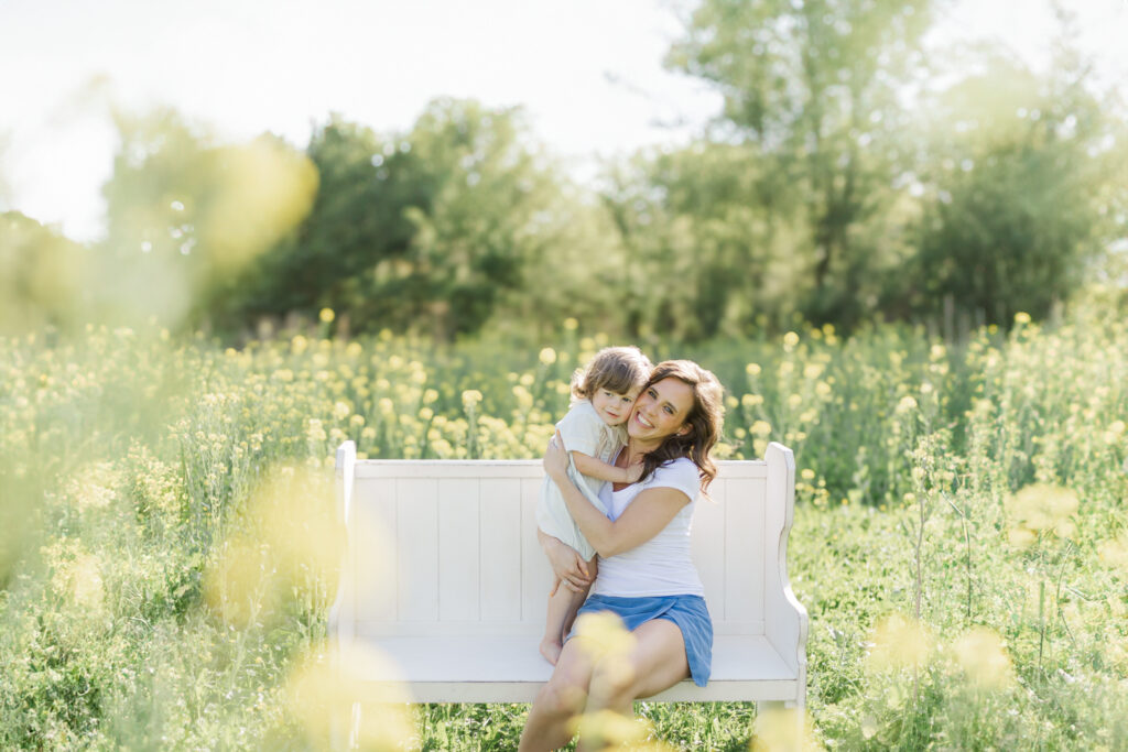 A smiling mother cuddling her toddler on a white bench surrounded by blooming yellow wildflowers, photographed outdoors in gentle spring light.