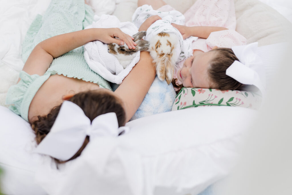 Two young girls lying together on white linens while gently holding baby bunnies, captured in soft natural light during a heirloom-style Easter mini photo session.