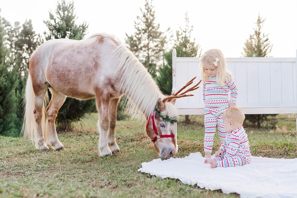 Two children wearing matching colorful Christmas pajamas next to Burt the Reindeer outdoors.
