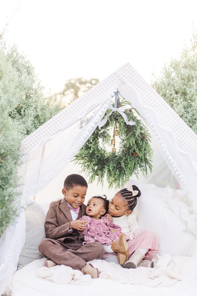 Children in delicate light-colored Christmas outfits photographed inside a cozy white holiday tent.