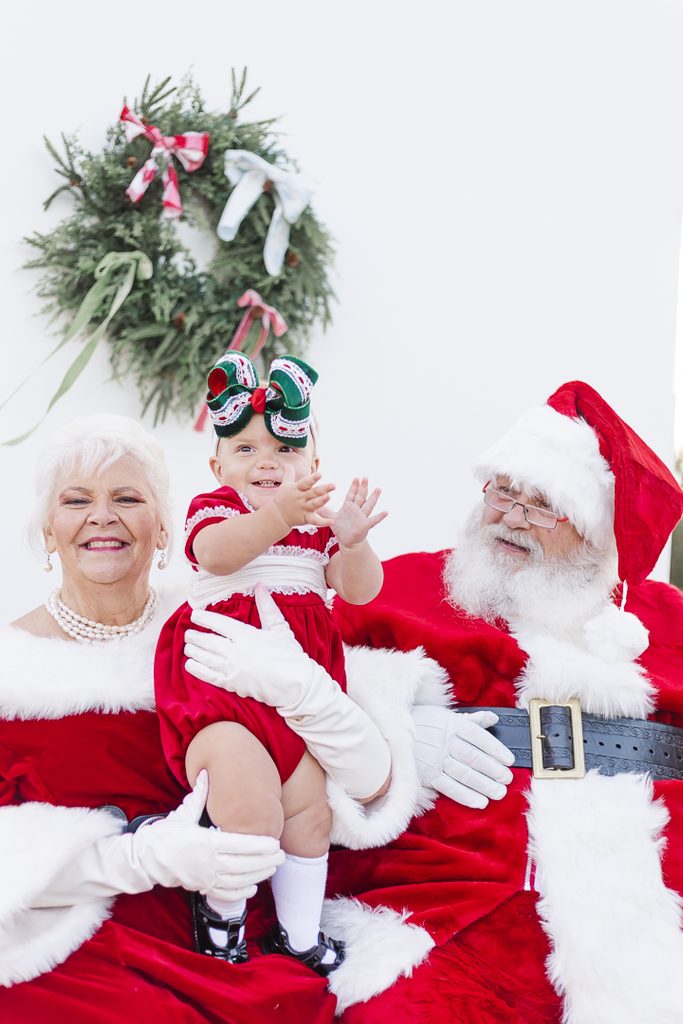 Mrs. Claus and Santa pose with a baby in a red and white Christmas outfit.