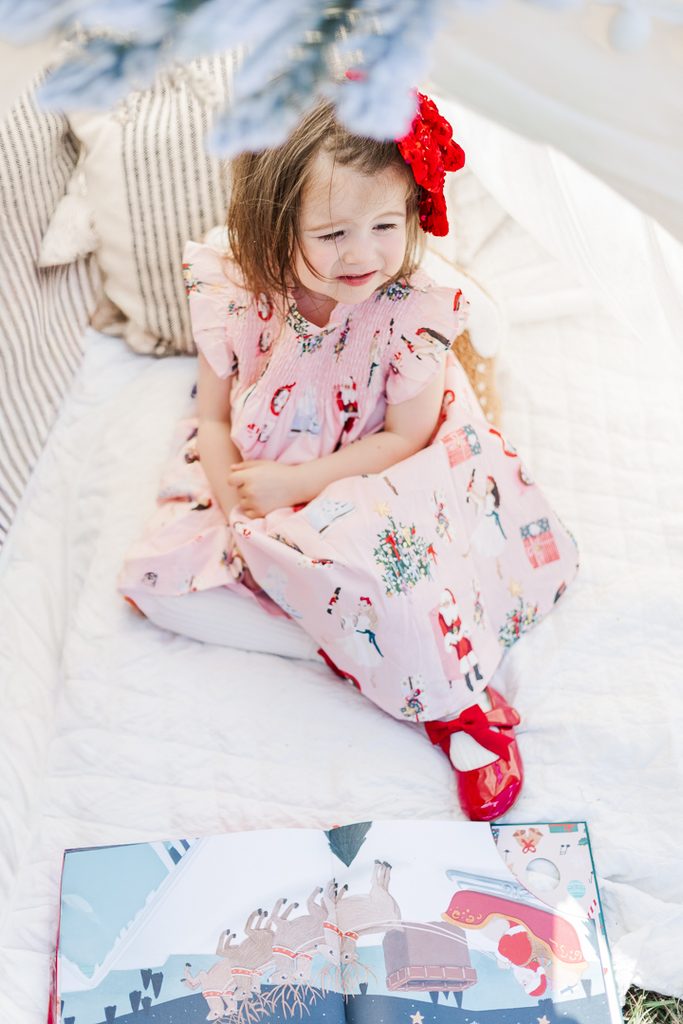 Young girl dressed in a patterned festive holiday dress sitting beside Christmas decorations.