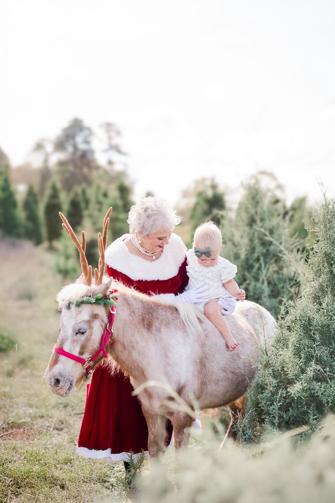 Mrs. Claus stands with a young child and Burt the Reindeer among evergreen trees.