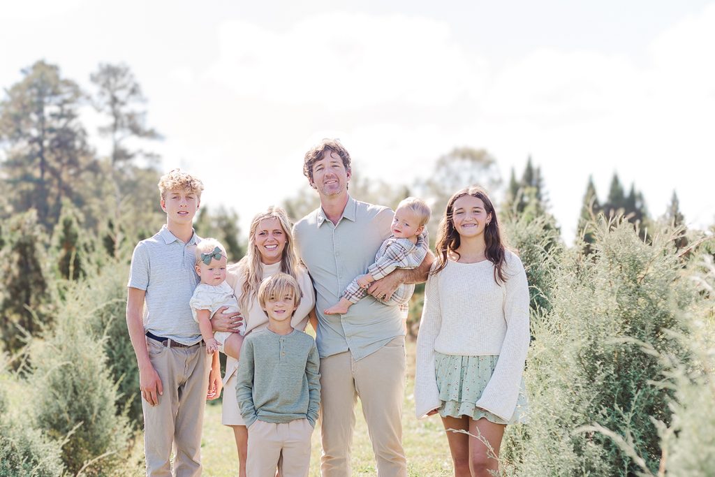 Family gathered outdoors among Christmas trees wearing coordinated soft neutral outfits.