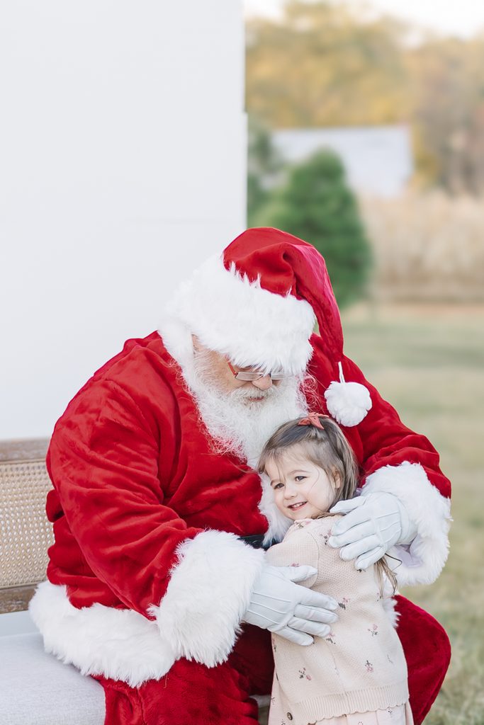 Santa Claus embraces a young girl in a classic red Christmas outfit during an outdoor holiday portrait.