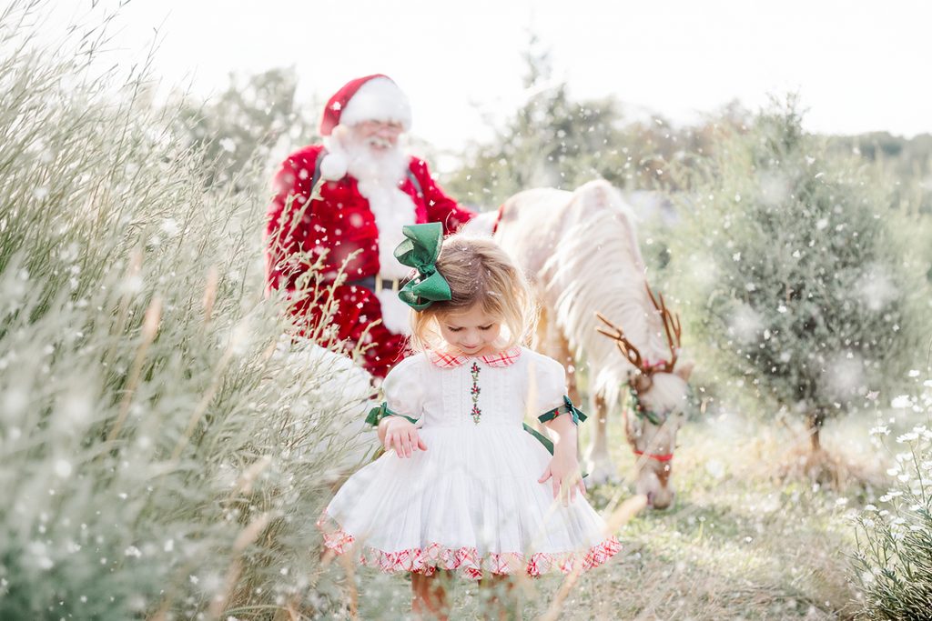 Little girl in a classic Christmas outfit walking among evergreen trees with Santa Claus and Burt the Reindeer.