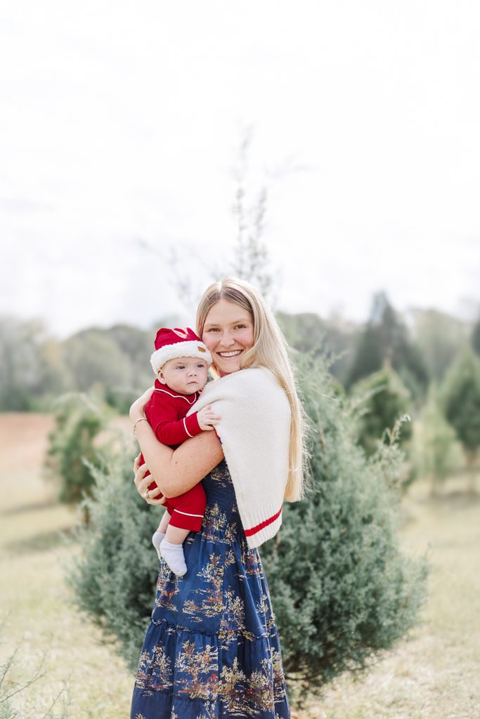 Mother holding a baby in a red Santa outfit among evergreen trees.