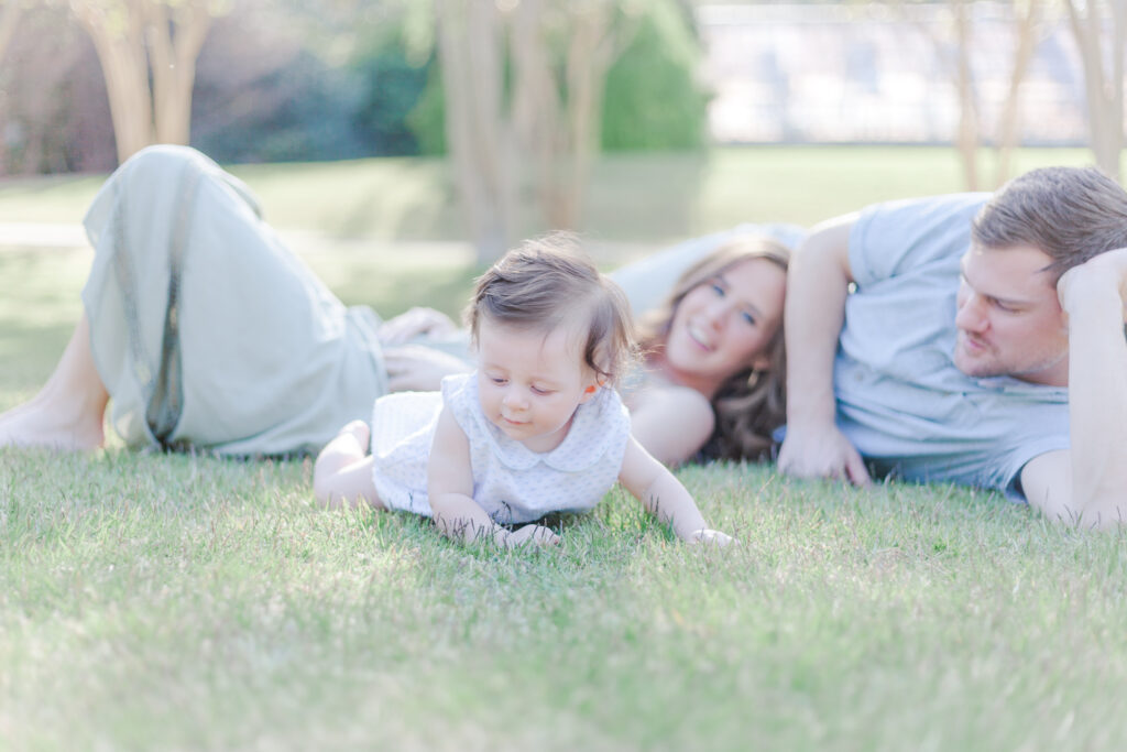 Family laying on grass with baby crawling toward the camera at the Birmingham Botanical Gardens during 6 Month Milestone Pictures with 5U Photography