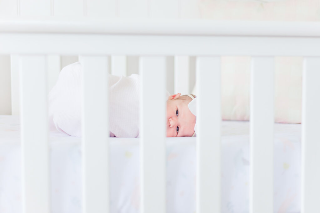 Newborn baby girl laying in cib on her side looking through crib slats with eyes open looking at camera during 5U Photography in-home, lifestyle newborn session