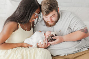 Mom and dad both holding baby girl looking down at baby sitting on bed smiling 