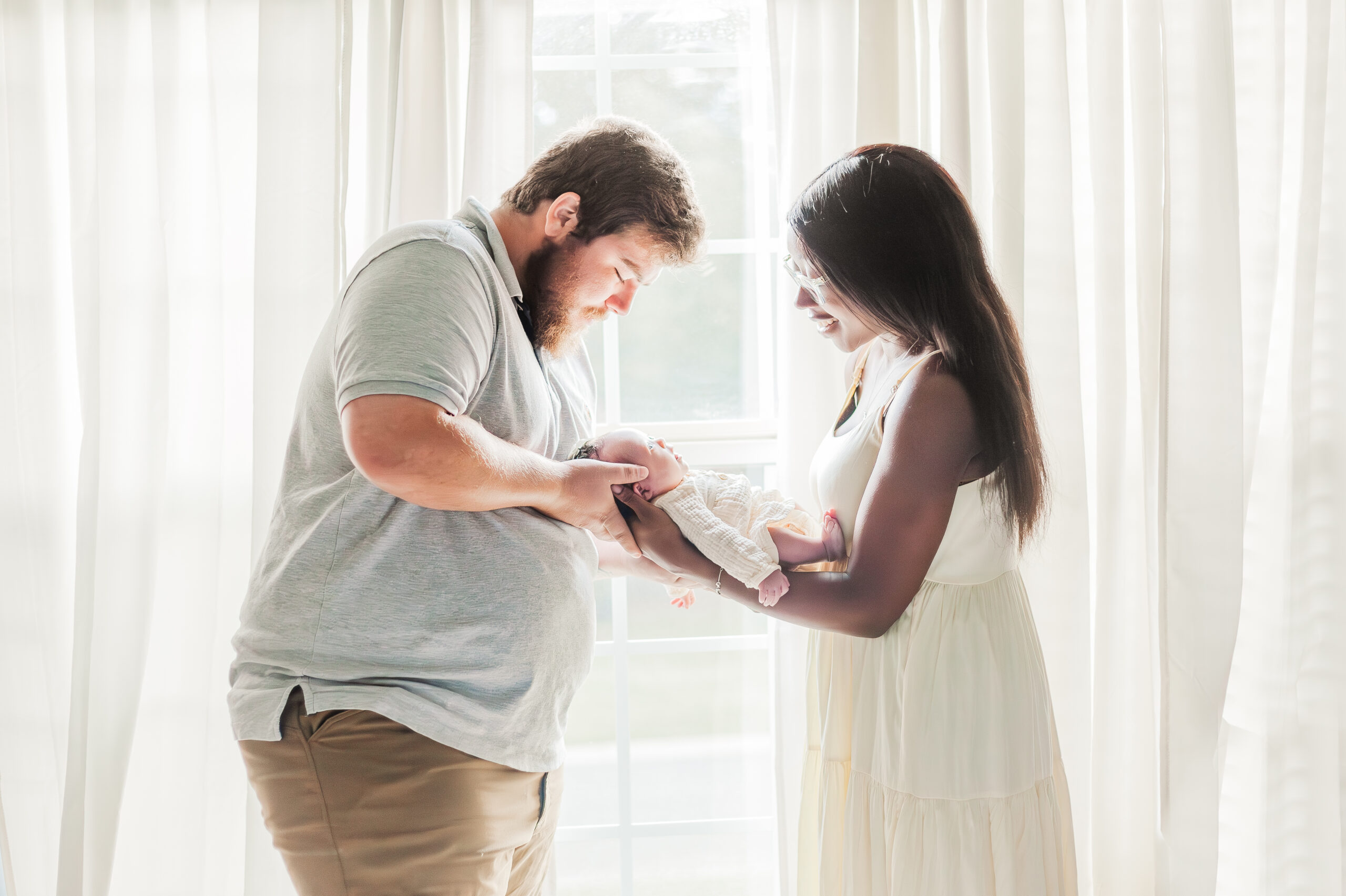 Mom and dad both holding baby girl standing in front of window looking down at baby during in-home newborn photo session