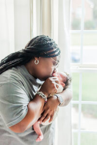 grandmother holding newborn baby girl kissing her hand standing in front of window during 5U Photography session