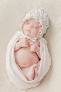 newborn baby girl wrapped in a pink blanket with flowered bonnet on laying with eyes closed 