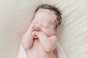 newborn baby girl laying with hands in front of face with white bow on during newborn session 