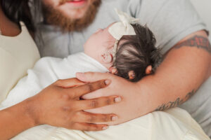 Close up of mom and dad's hands holding baby girl with white bow during in-home newborn session