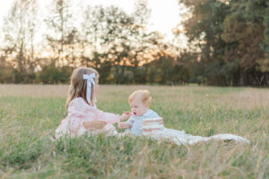 Little girl sitting with brother on blanket feeding piece of cake during one year milestone and smash cake session