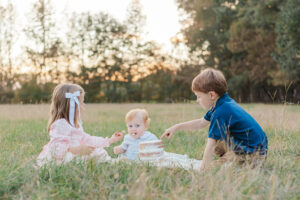 1 year old red headed little boy sitting on blanket with brother and sister during 1 year milestone session eating smash cake 