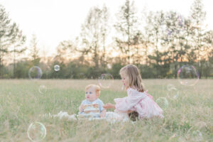 1 year old little boy sitting with sister on blanket with smash cake looking at bubbles during 1 year milestone and smash cake session