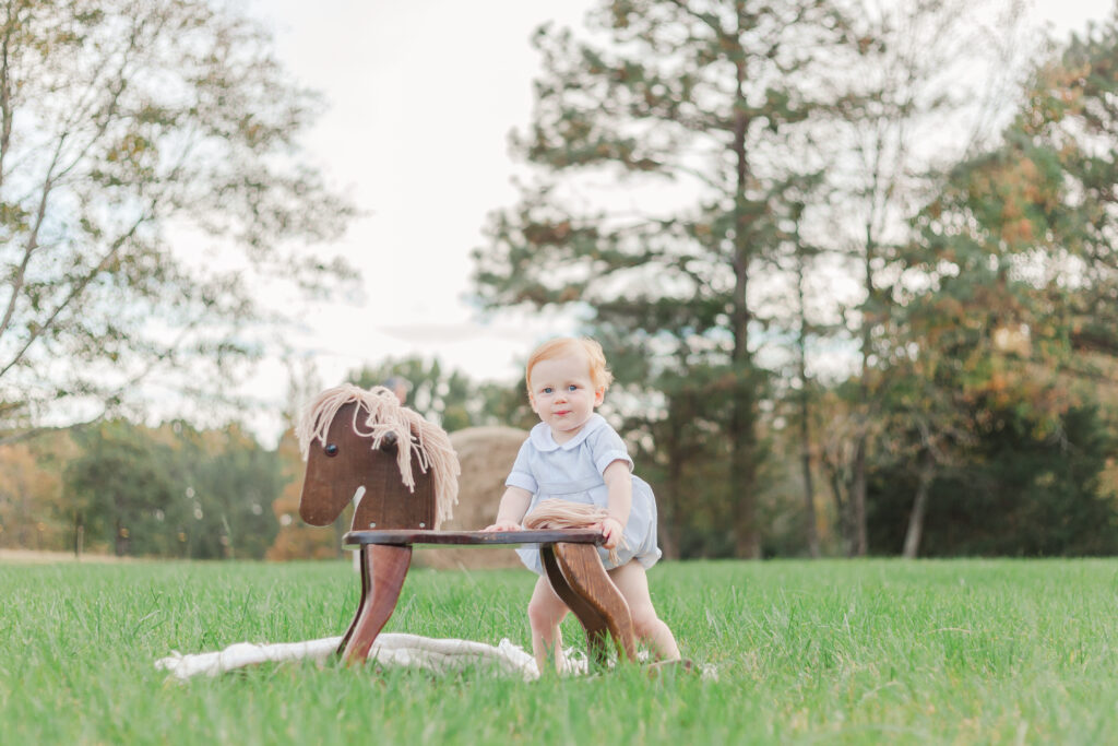 1 year old little boy standing behind rocking horse in a field smiling at camera during 1 year milestone photography session with 5U Photography 