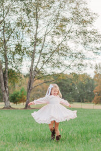 little girl in pink gingham dress with white boy spinning in field smiling down during brother's 1 year milestone photo session